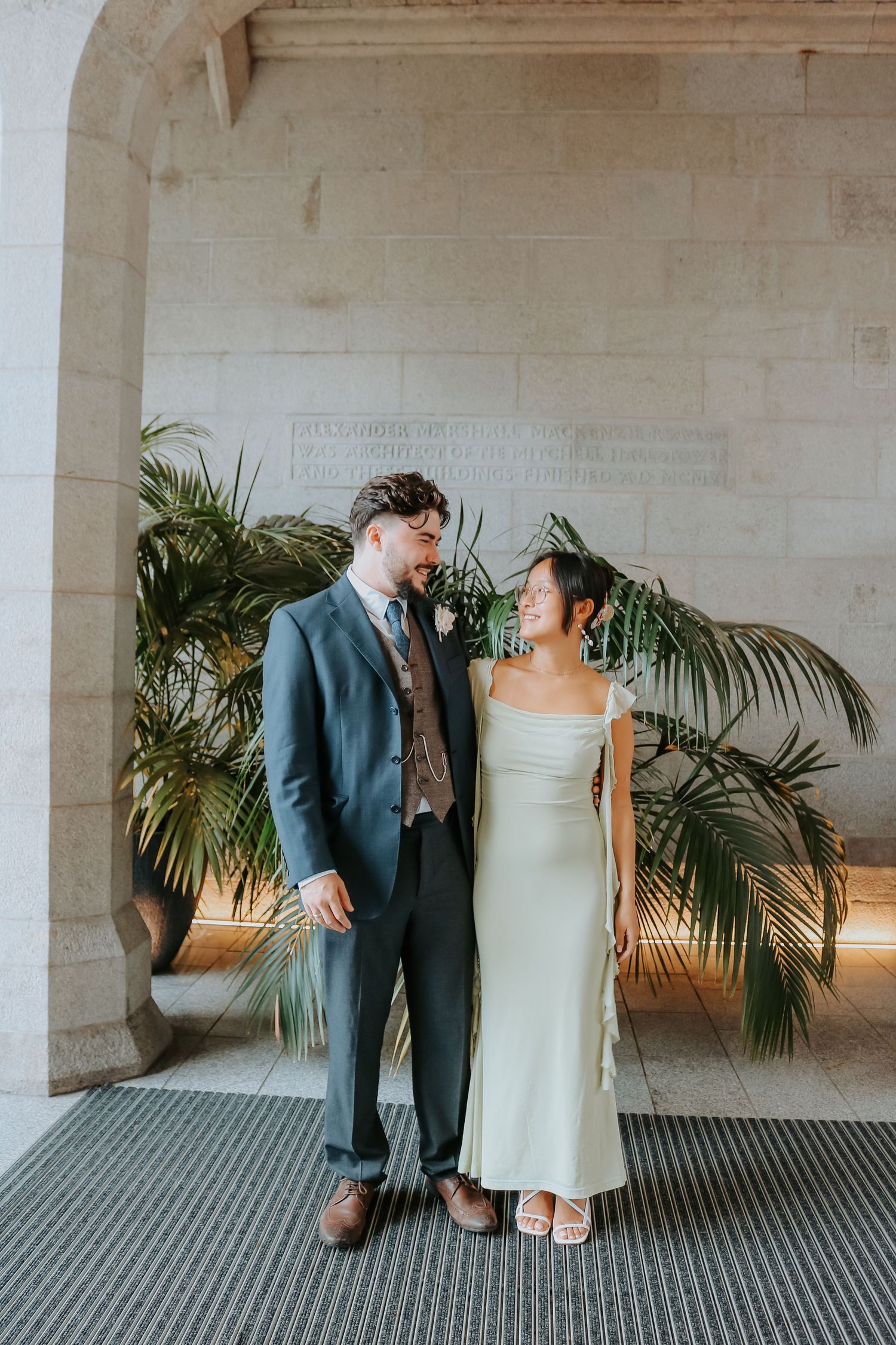 A couple shares a romantic moment under an archway with palm plants, the bride wearing a sleek white gown.