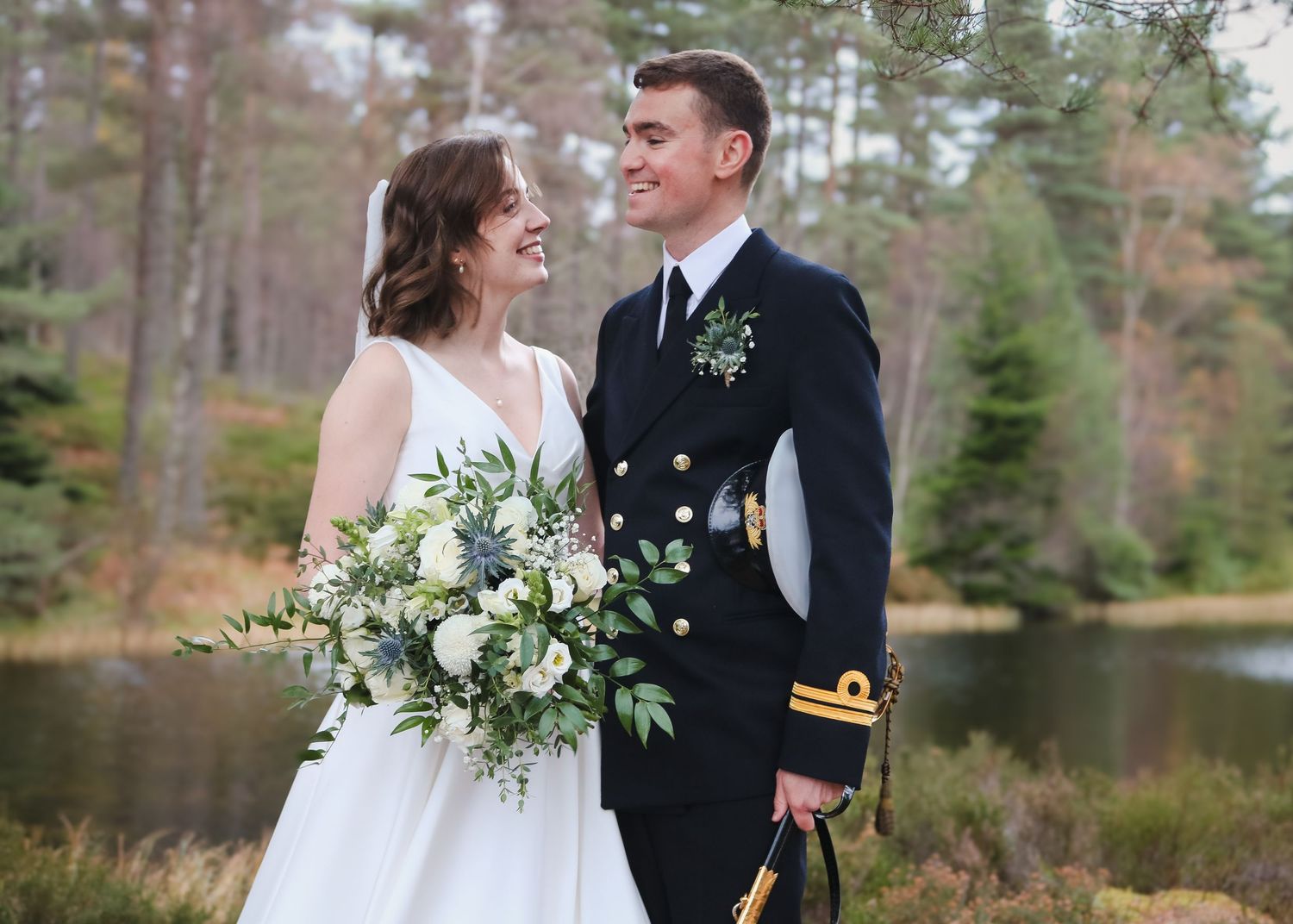 A newlywed couple shares a joyful moment by a serene lake with pine trees in the background.
