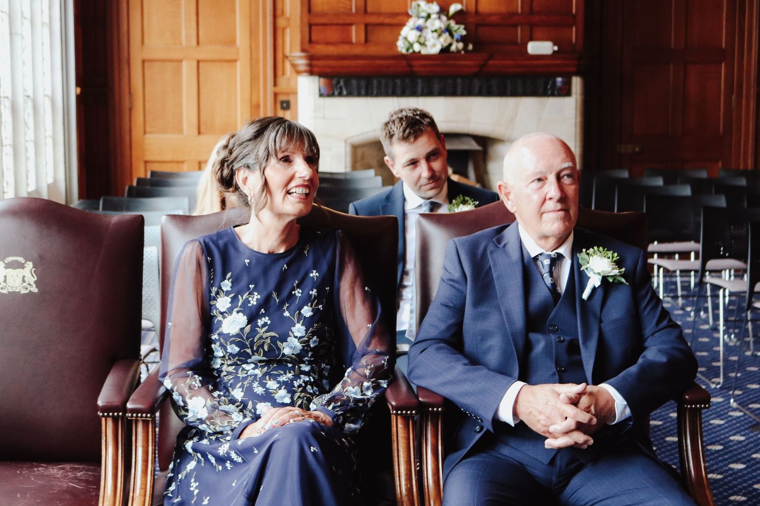 Three people sit next to each other on brown upholstered chairs at a formal event with glassware visible in the background.