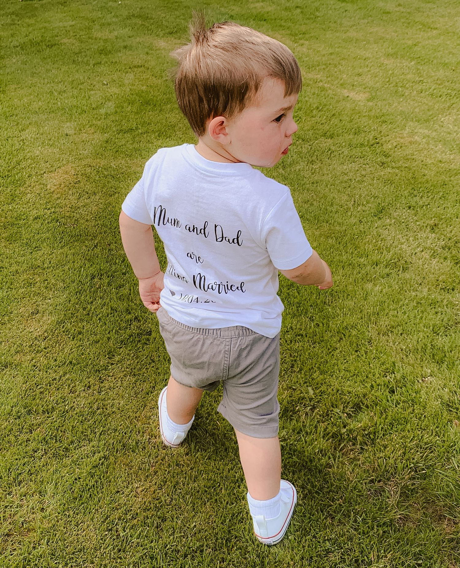 A toddler wearing a white t-shirt and gray shorts walking on grass outdoors.