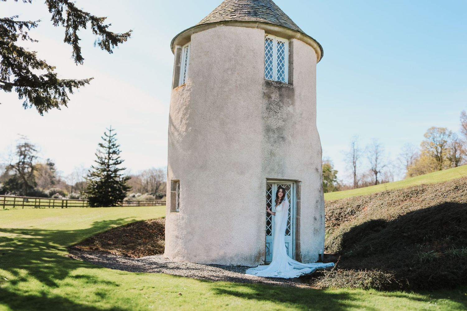 A cylindrical white tower with a domed roof and blue door sits on a grassy hill against a clear sky.