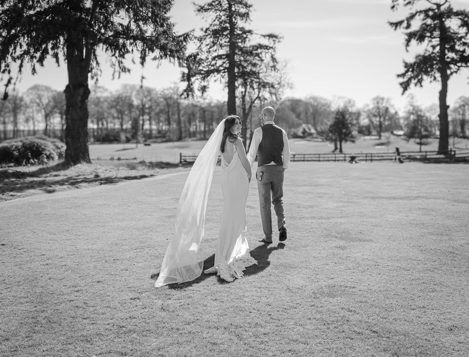 A black and white vintage photograph shows a bride and groom standing on a lawn surrounded by tall trees.