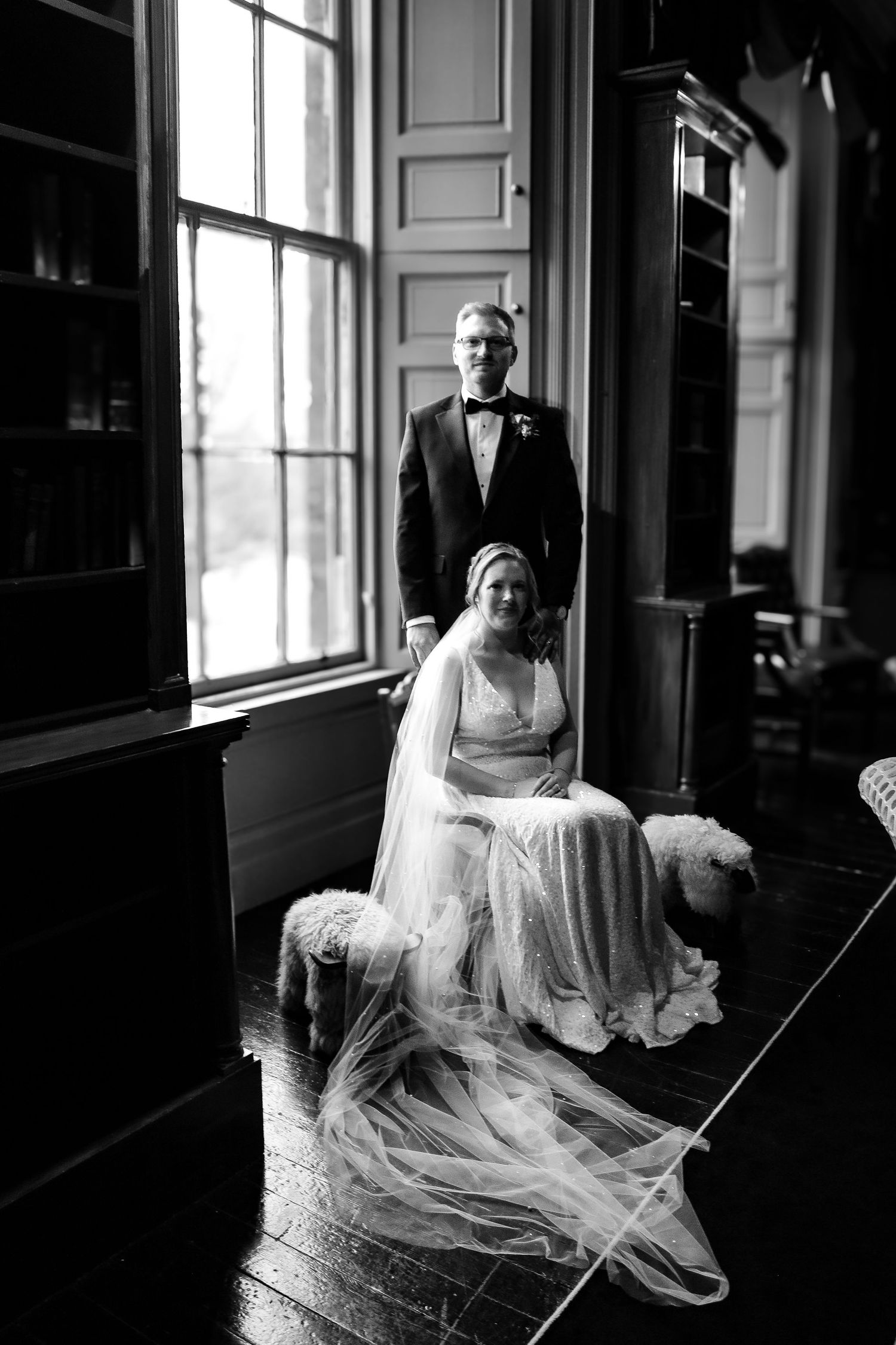 Black and white wedding portrait of a couple in an elegant room with tall windows and flowing veil details.