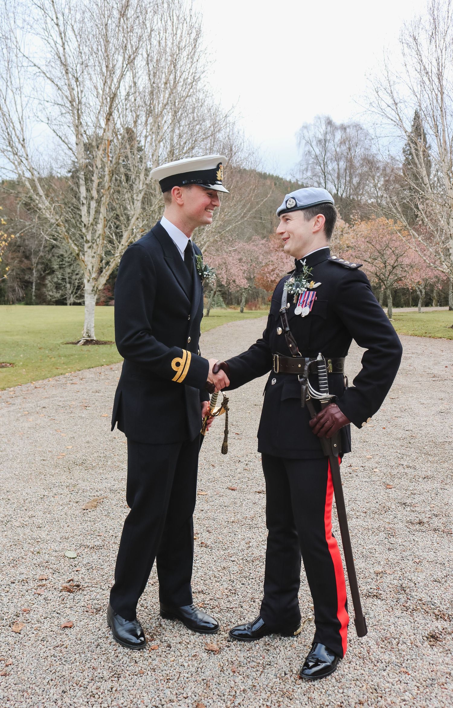Two naval officers in formal military dress uniforms shake hands on a gravel path surrounded by bare winter trees.