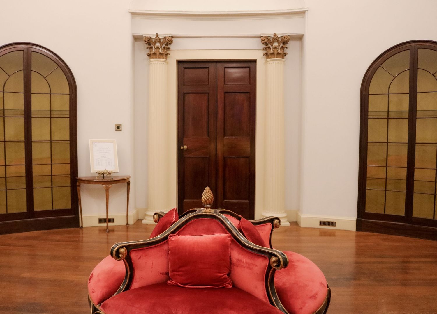 An elegant room interior with arched windows, dark wooden door, and a plush red velvet sofa in the foreground.