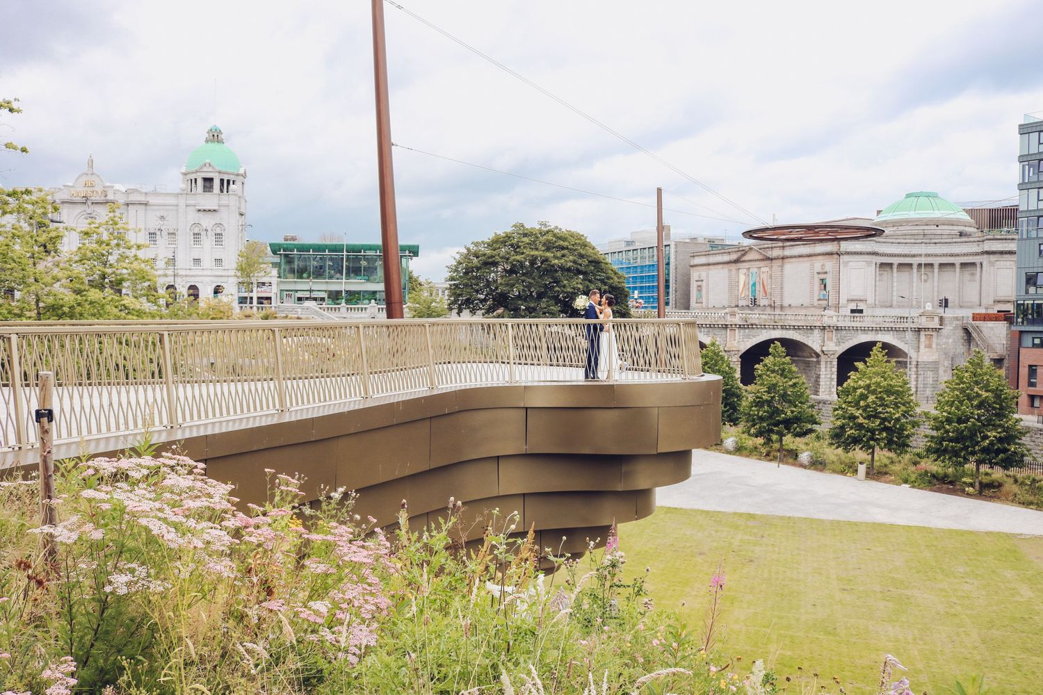 A small pedestrian bridge with ornate railings spans across a grassy area near historic buildings on a cloudy day.