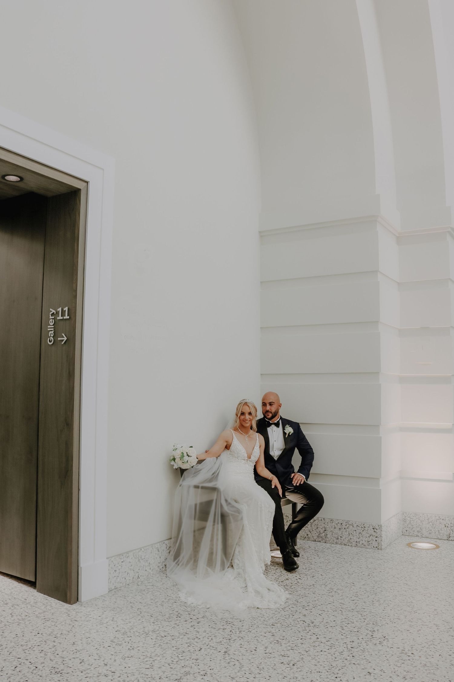 A couple in wedding attire sitting beside an elevator in a modern white hallway with terrazzo flooring.