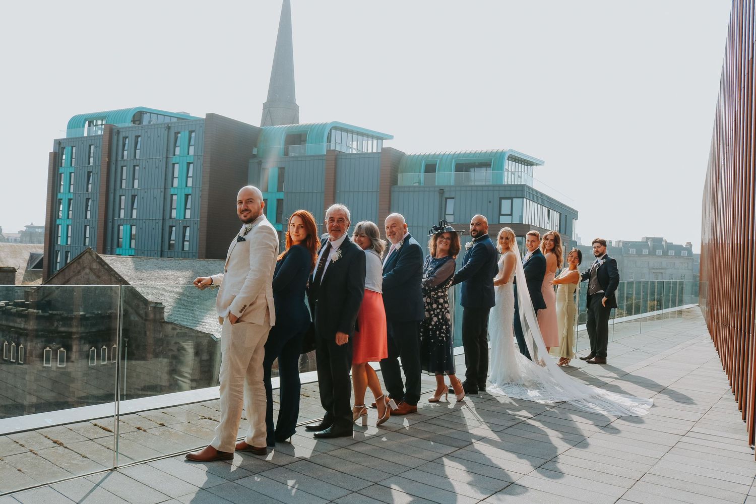 A wedding party poses together on a rooftop with modern buildings and a church spire in the background.
