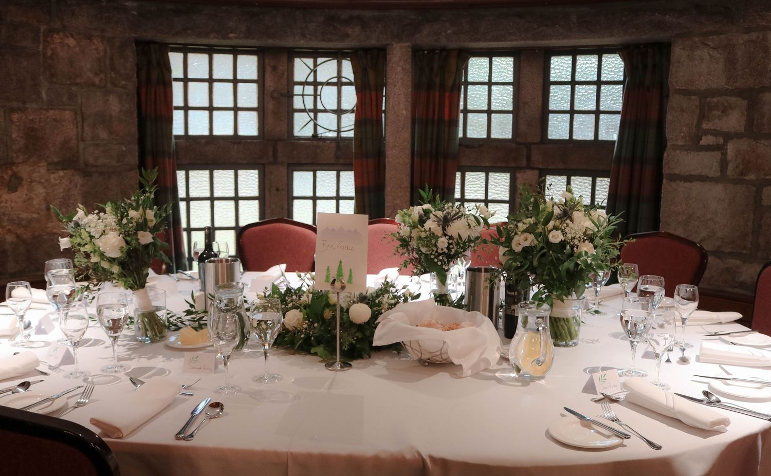 Round table with elegant white floral centerpieces and formal place settings in rustic stone venue with window views.