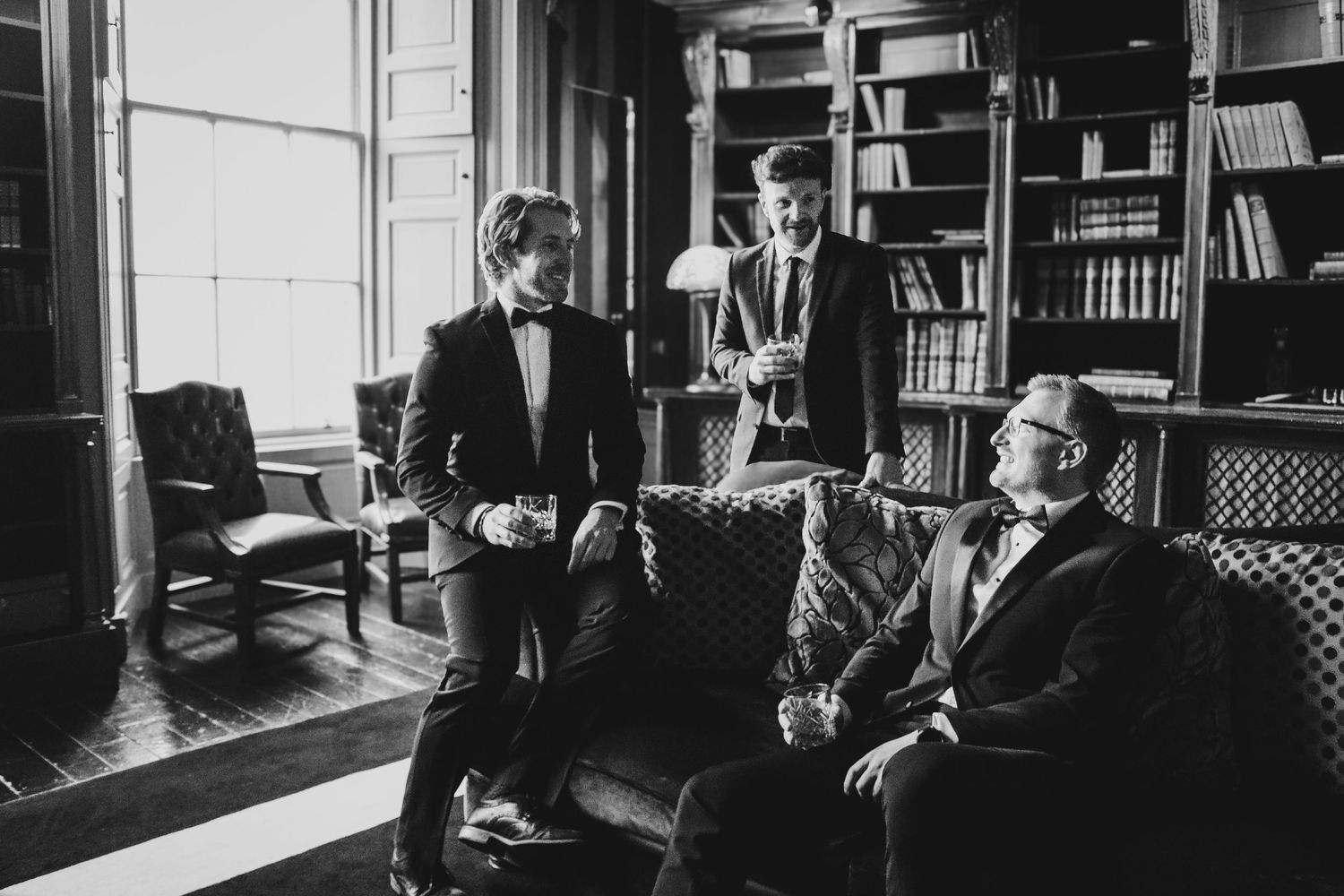 Three people gather in a vintage library setting with bookshelves and period furniture in black and white.