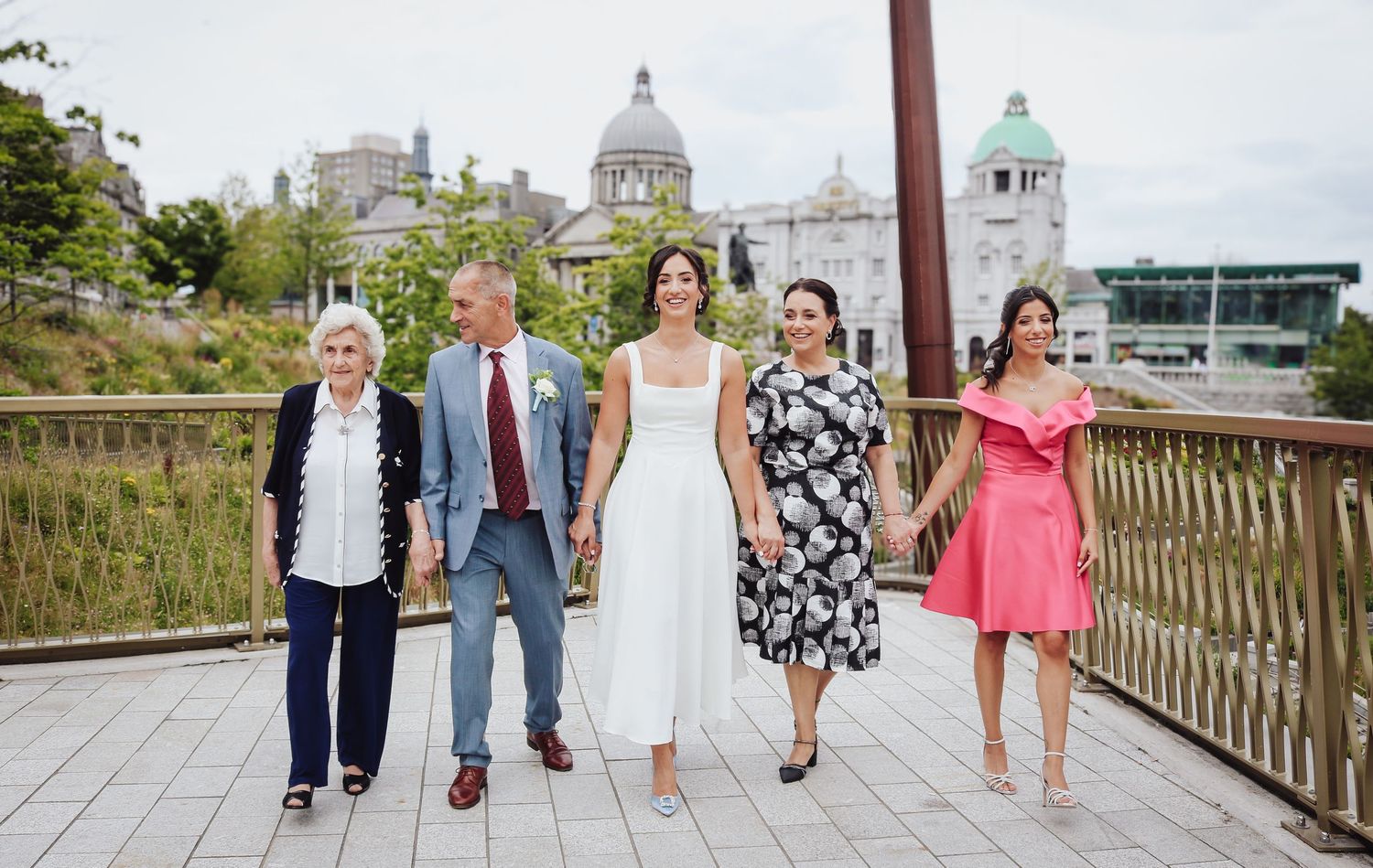A cheerful group strolls along a riverside walkway with historic domed buildings in the background in Liverpool.