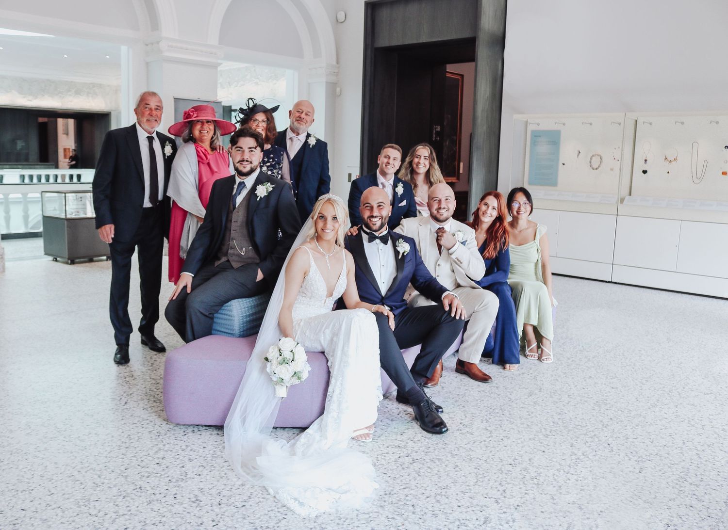 A wedding party poses together on seating in a bright modern lobby.