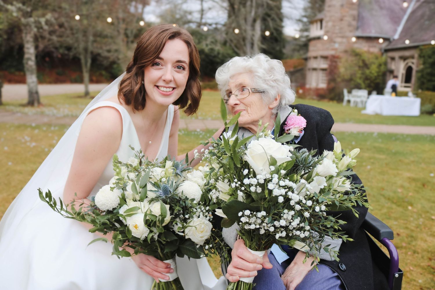 Two people share a heartwarming moment while holding white and green flower bouquets outdoors on a grassy lawn.