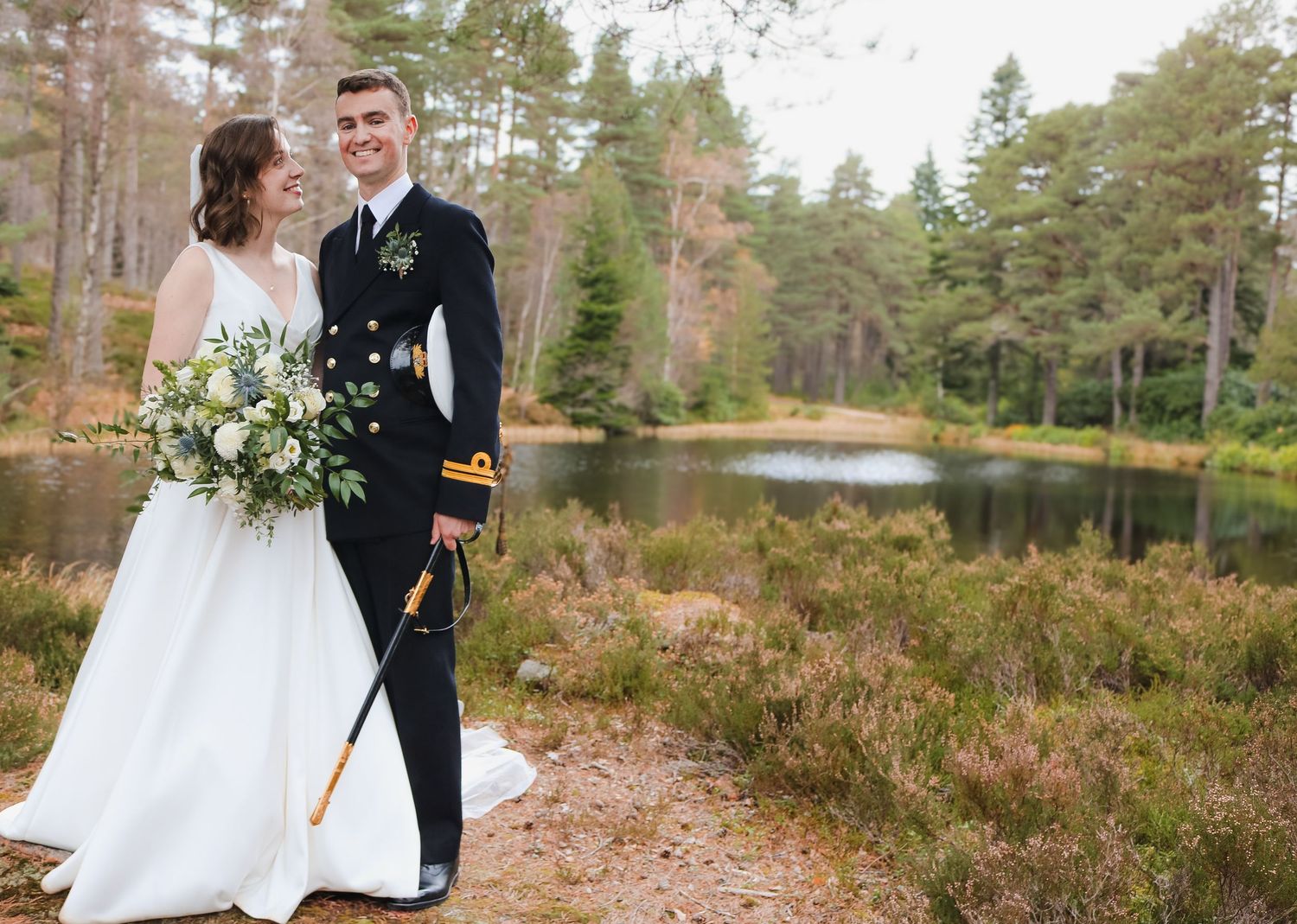 A wedding couple stands near a serene pond in the woods, with fall foliage and evergreen trees in the background.