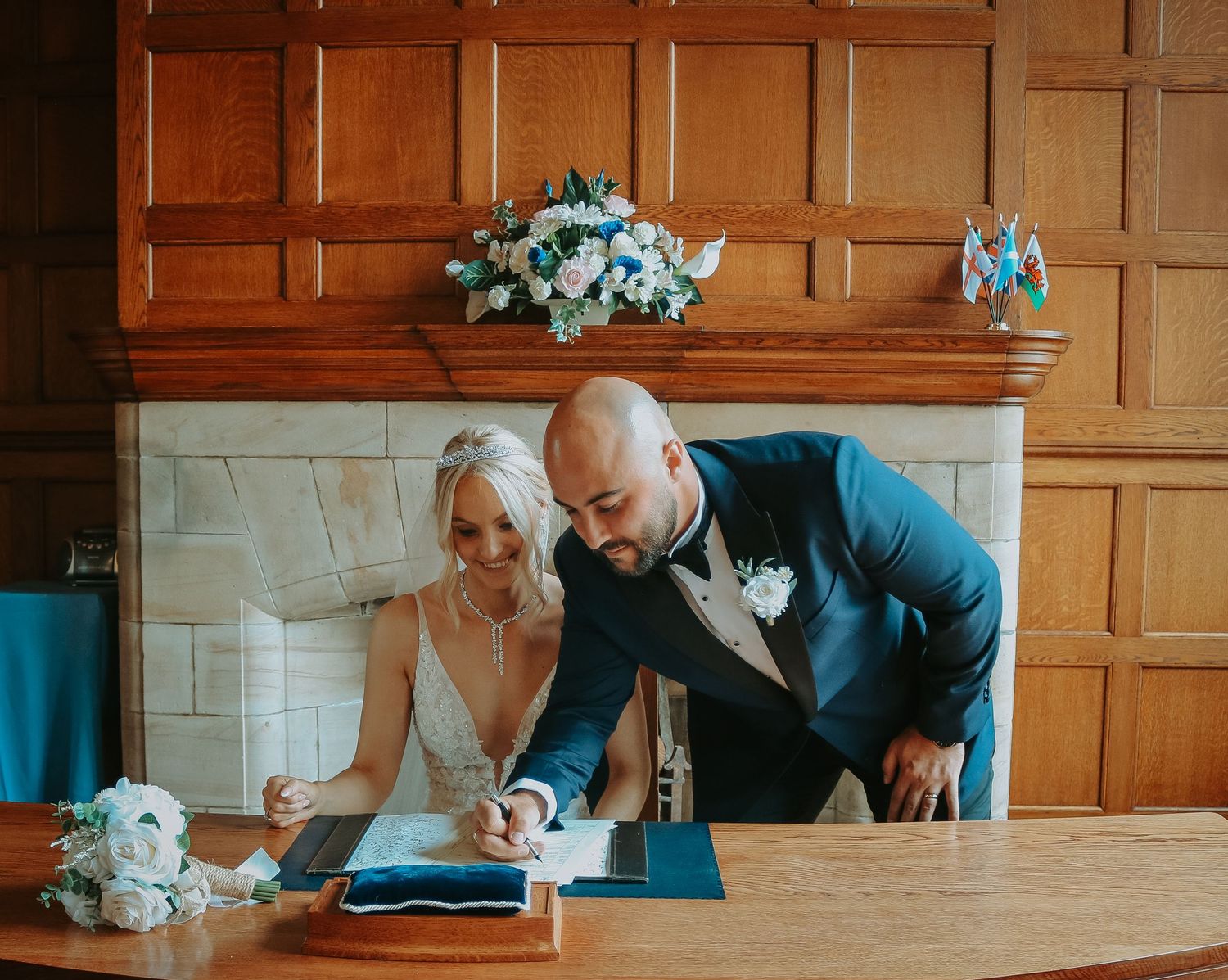 Couple in wedding attire lean over a table while signing documents in a room with wood-paneled walls.