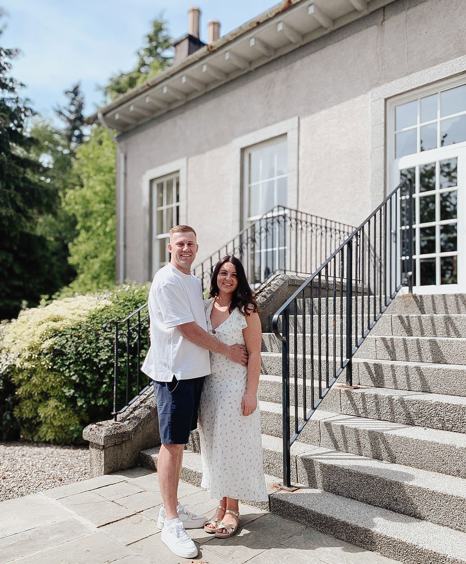 A couple stands on stone steps outside a gray stucco house with large windows and iron railings.