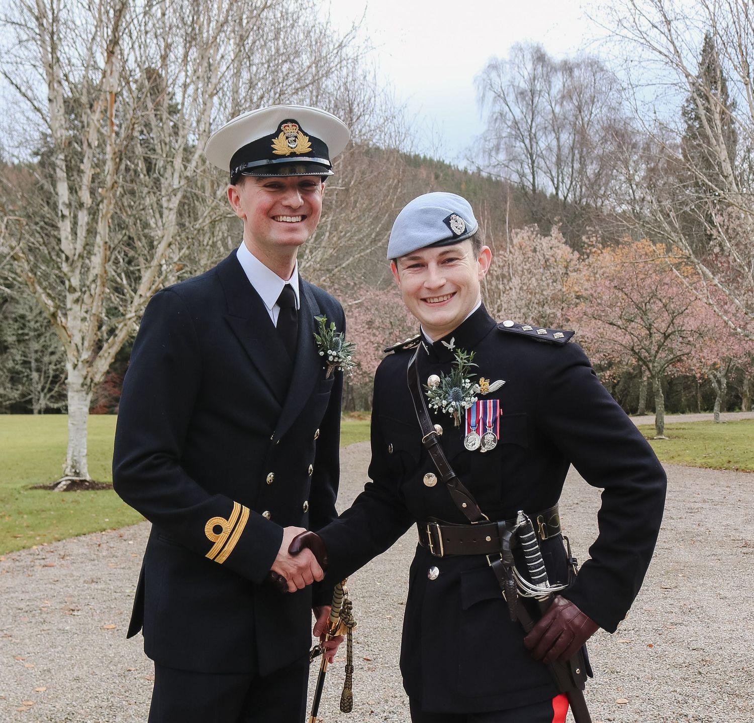 Two uniformed military service members shaking hands outdoors near birch trees on a cloudy day.