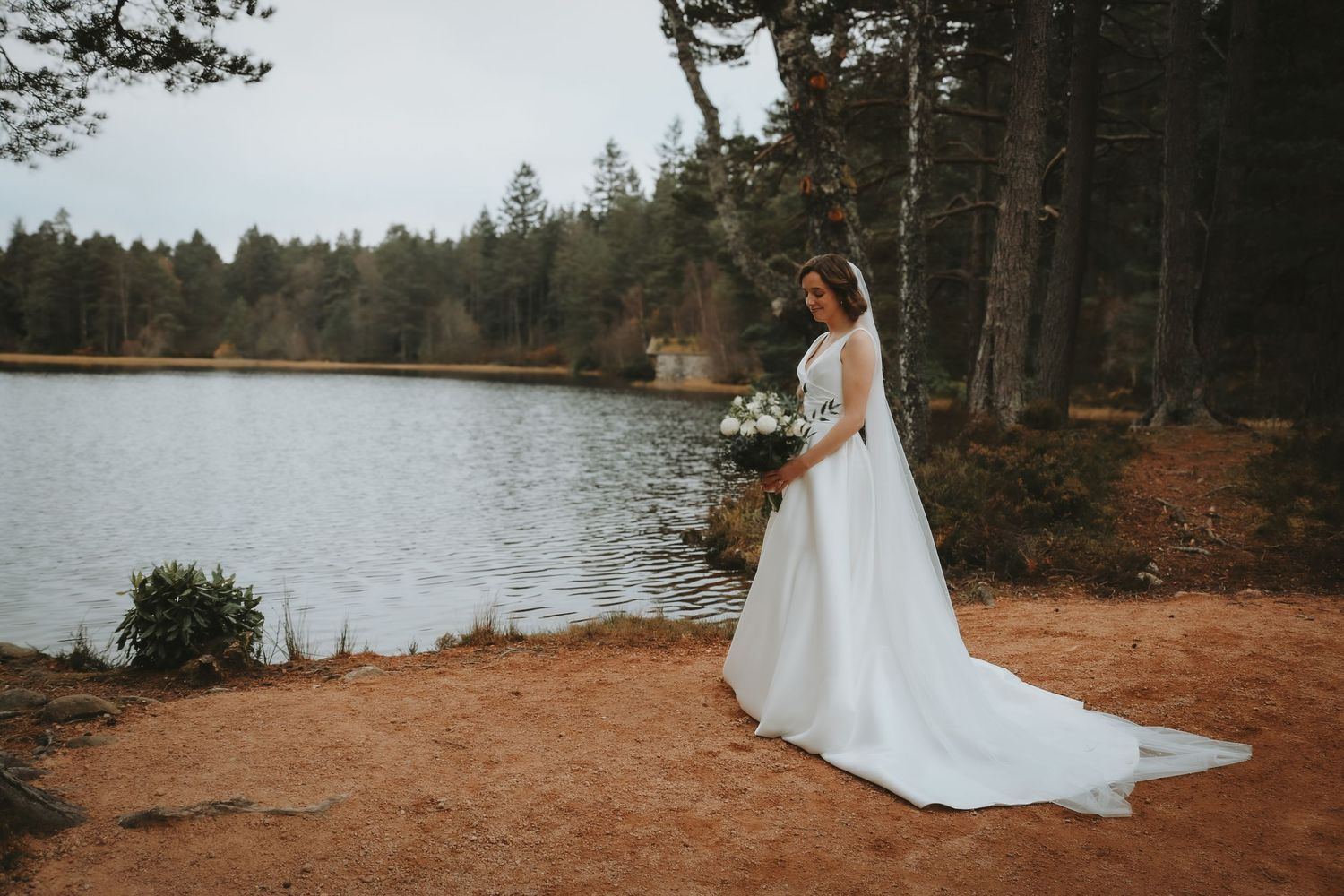 A person in a flowing white wedding dress stands on the rustic shoreline of a serene lake surrounded by evergreen forest.