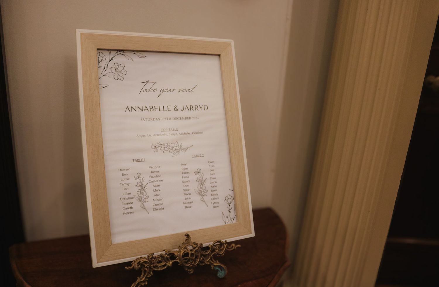 A framed wedding seating chart displayed on a wooden surface with elegant script text and floral details.