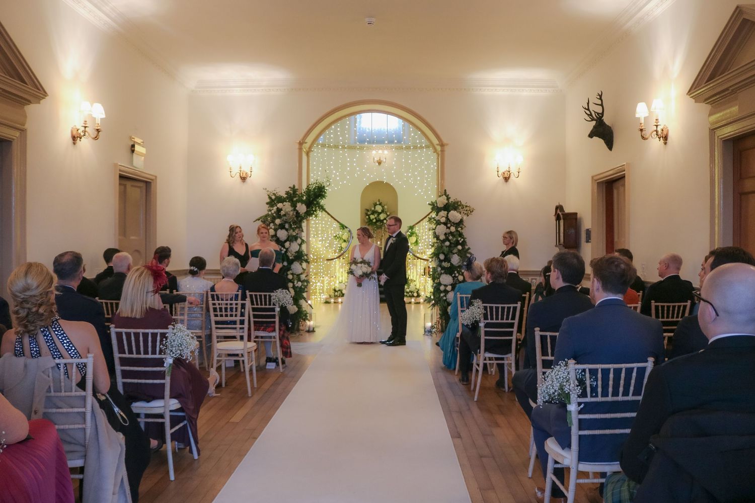 Wedding ceremony with white carpet aisle and floral arch in elegant venue with guests seated in Chiavari chairs.