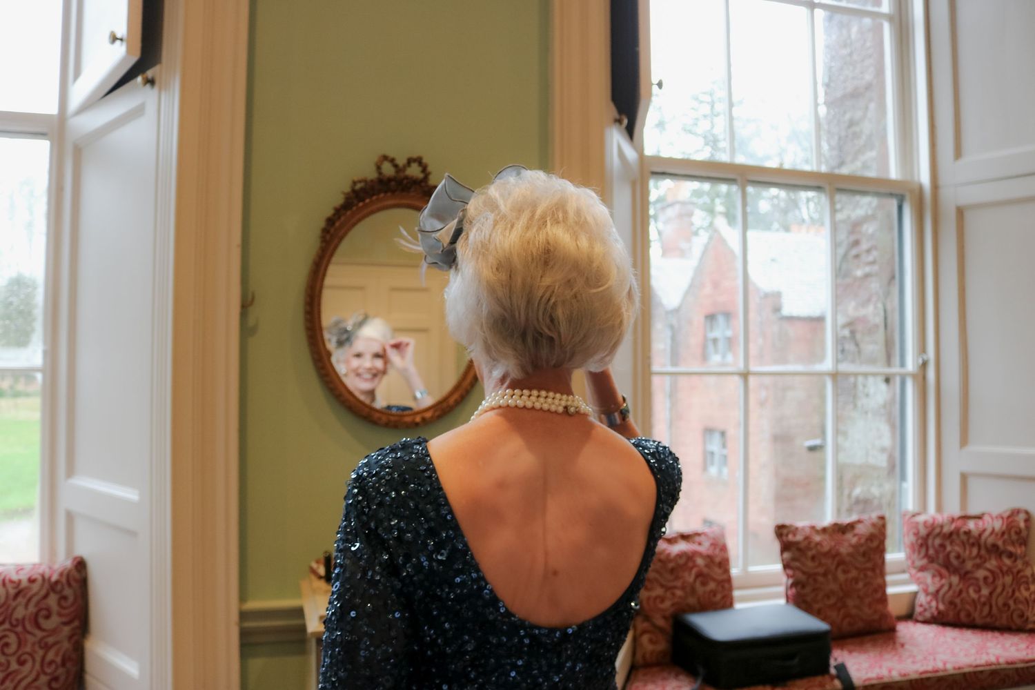 Woman in sparkly black evening dress gazes out bright bay window with round mirror on wall beside her.