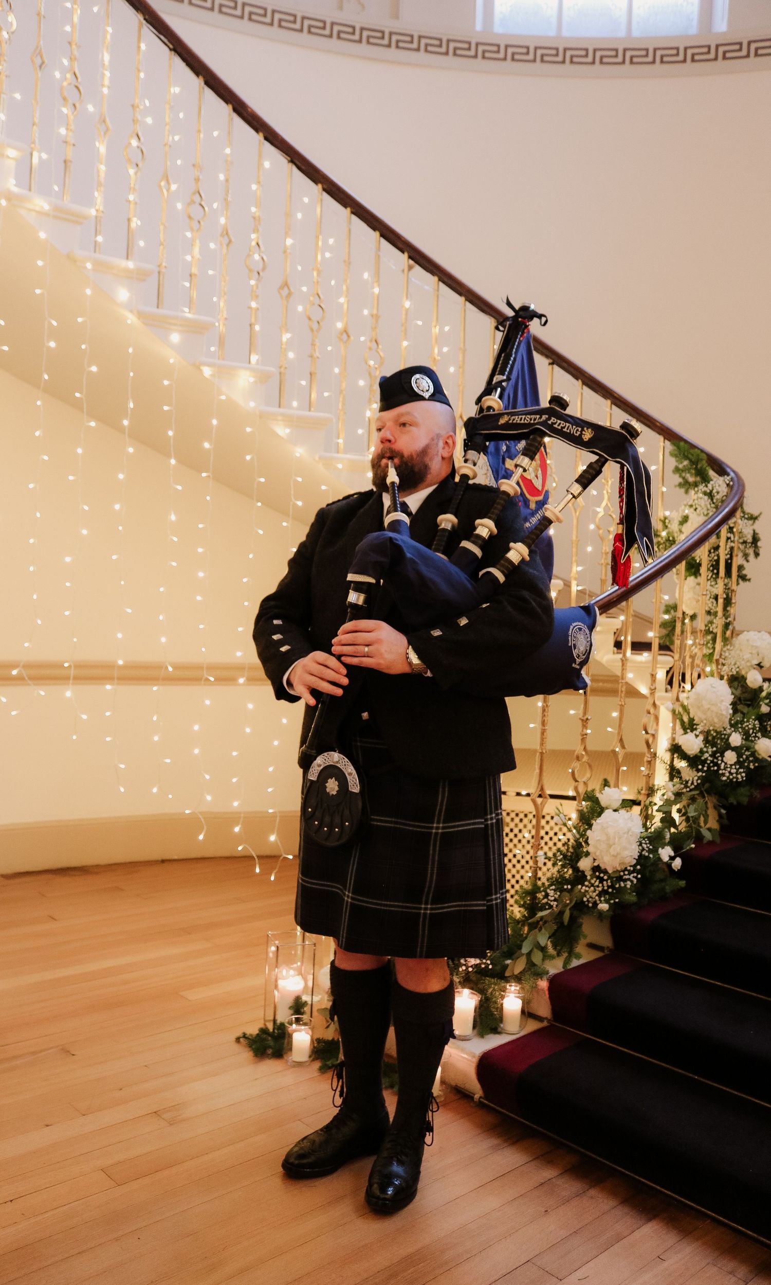 A bagpiper in traditional Scottish attire performs next to a decorated staircase with twinkling lights and candles.