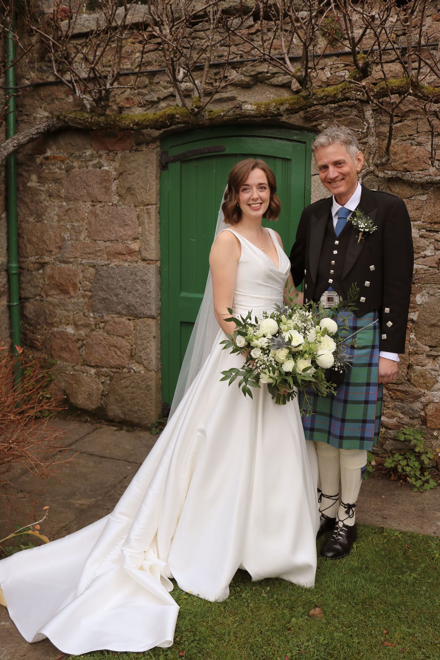 Couple in traditional Scottish wedding attire pose by green door against stone wall with bride holding white bouquet.