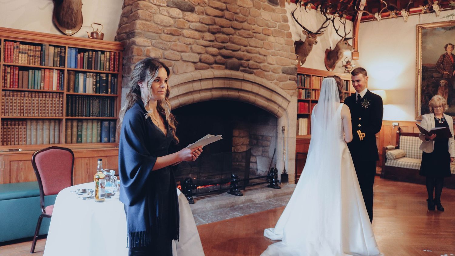 A wedding ceremony takes place in a rustic room with stone fireplace, wood beams, and bookshelves.