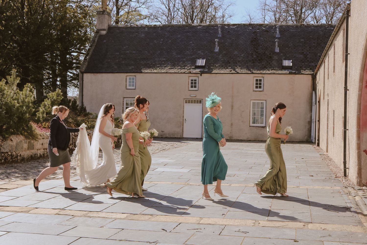 A playful group in formal attire skips along a stone path by a beige stone building on a sunny day.