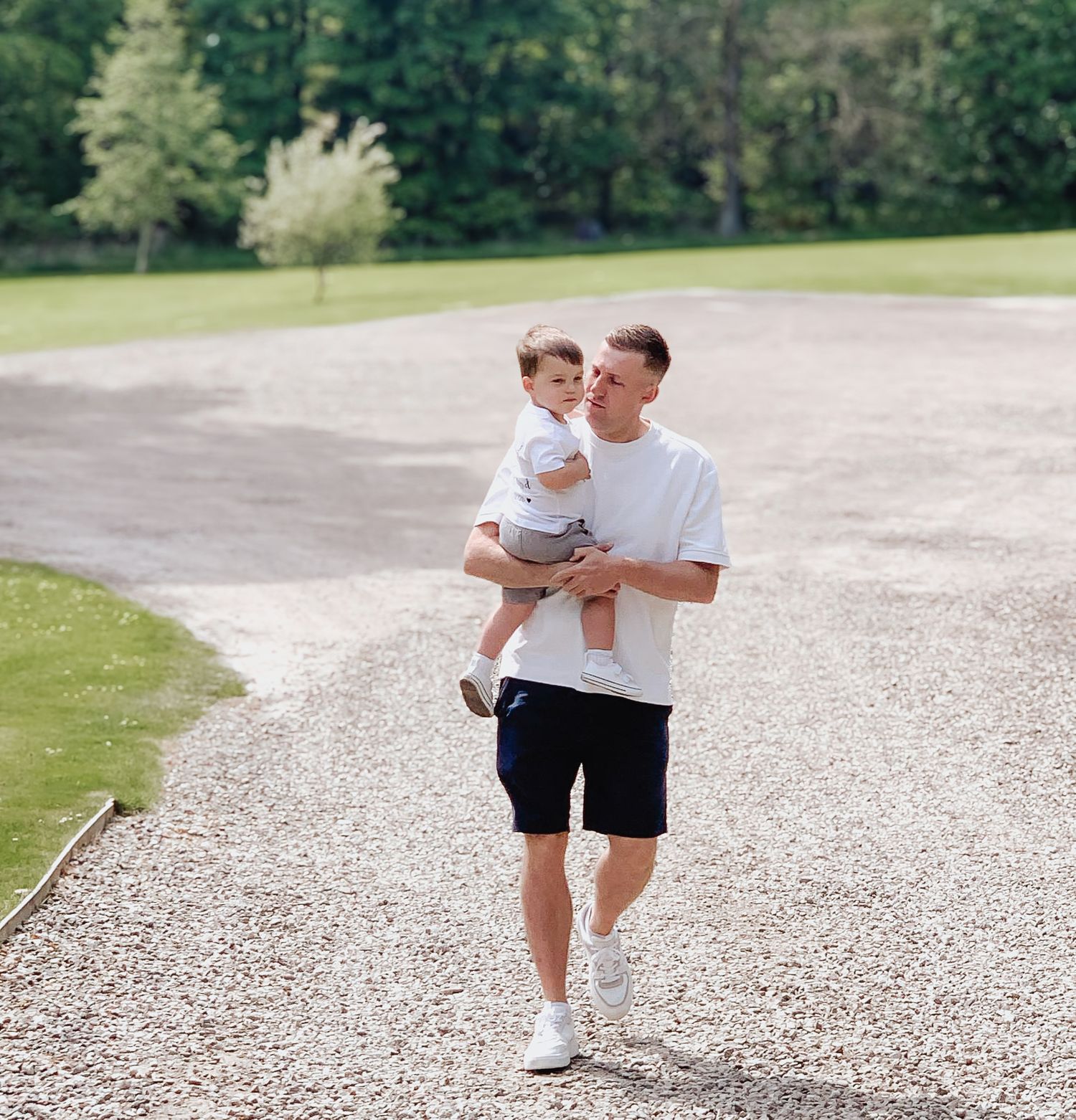A parent carries a small child while walking along a gravel path bordered by green lawn and trees.