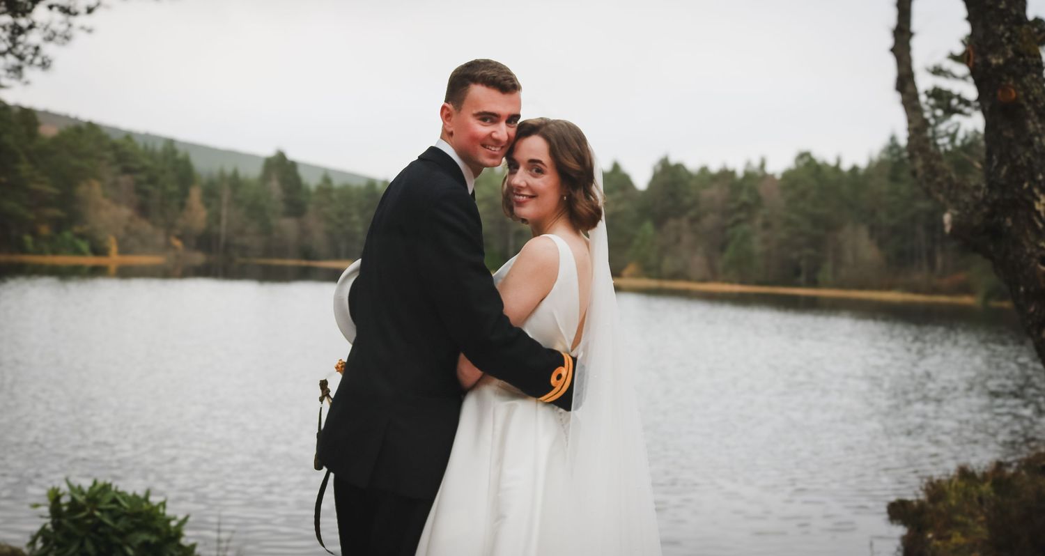 A romantic newlywed couple embrace by a serene mountain lake surrounded by autumn trees in a scenic outdoor setting.