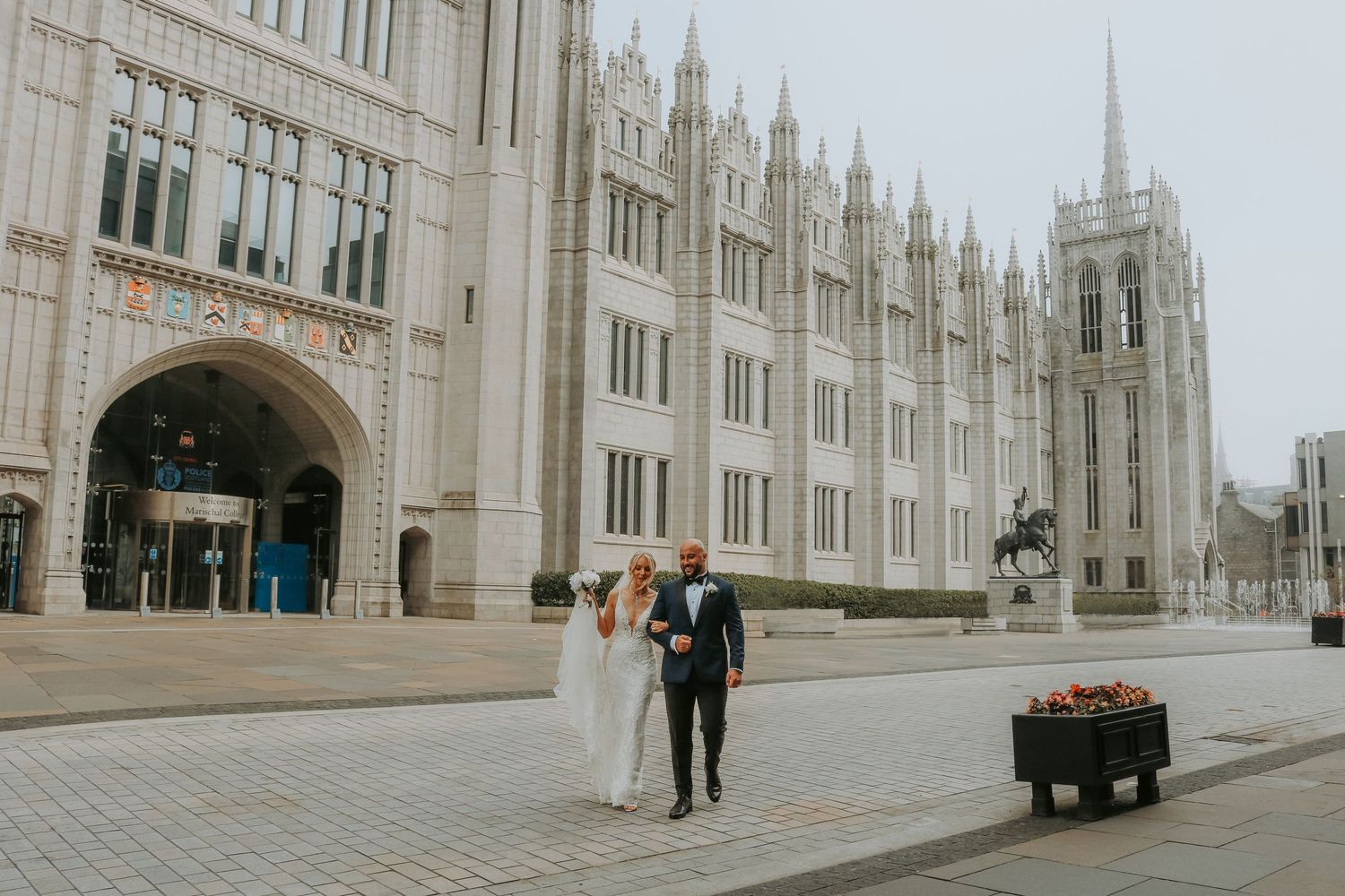 A couple stands outside the ornate gothic architecture of Marischal College in Aberdeen on an overcast day.