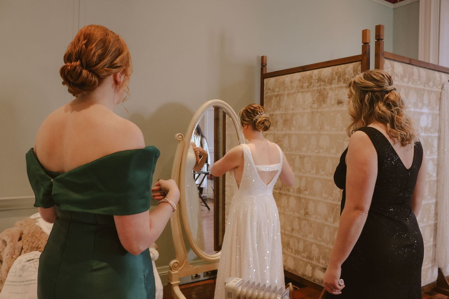 A person in an emerald green dress stands beside someone in a white gown during wedding preparations in a bridal suite.