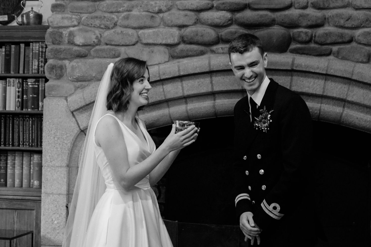 Black and white photo of a bride and groom sharing a laugh together against stone archway during wedding celebration.
