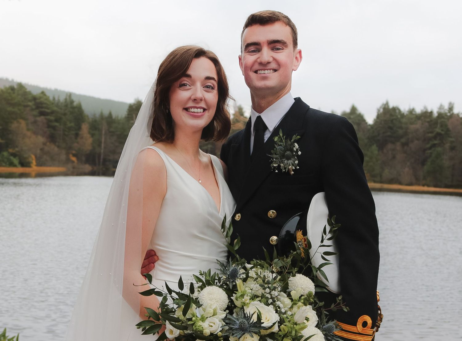 Newlyweds pose together by a tranquil lake with white floral bouquet and traditional wedding attire.