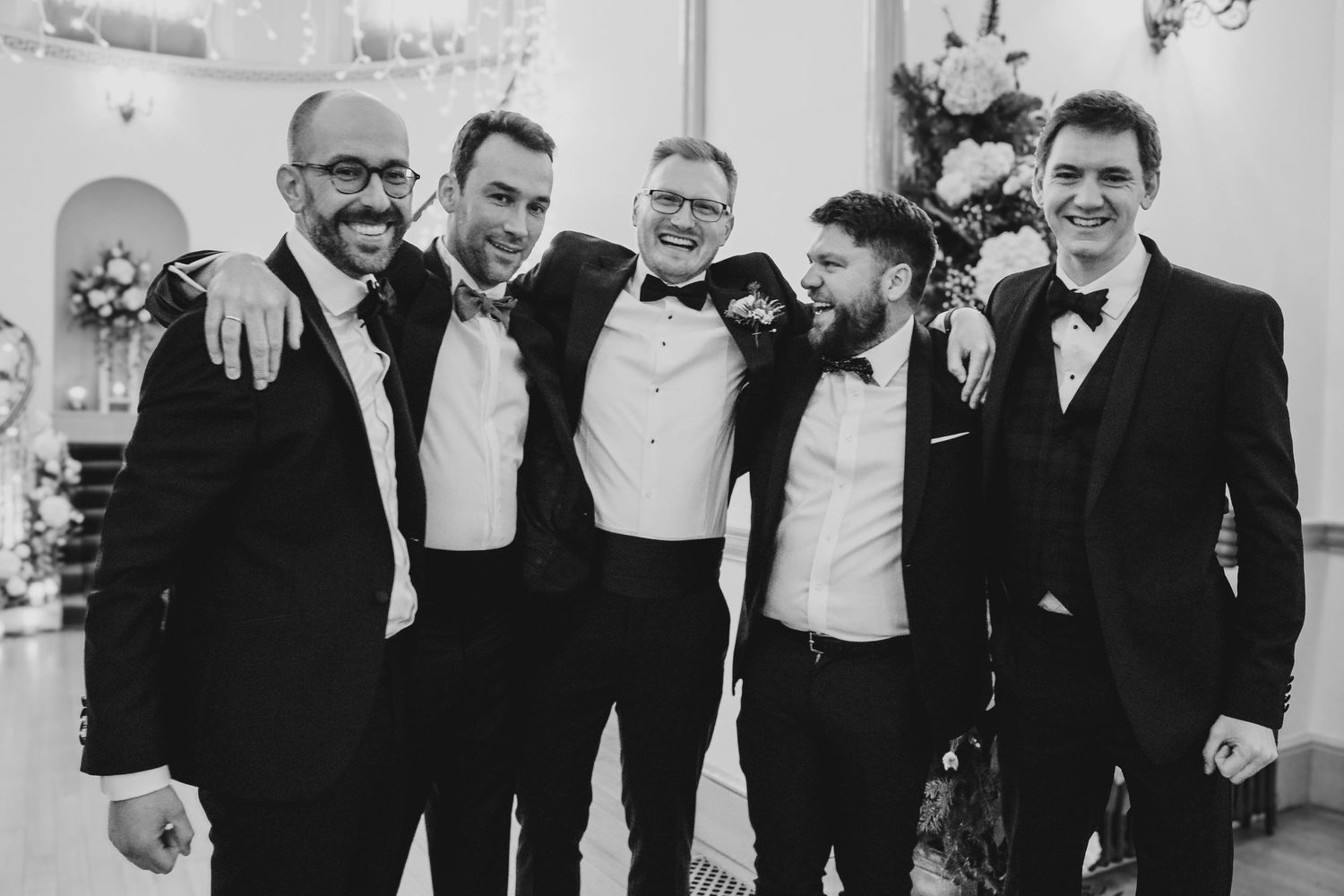 Groomsmen in formal attire smile together at a wedding celebration in black and white photography.