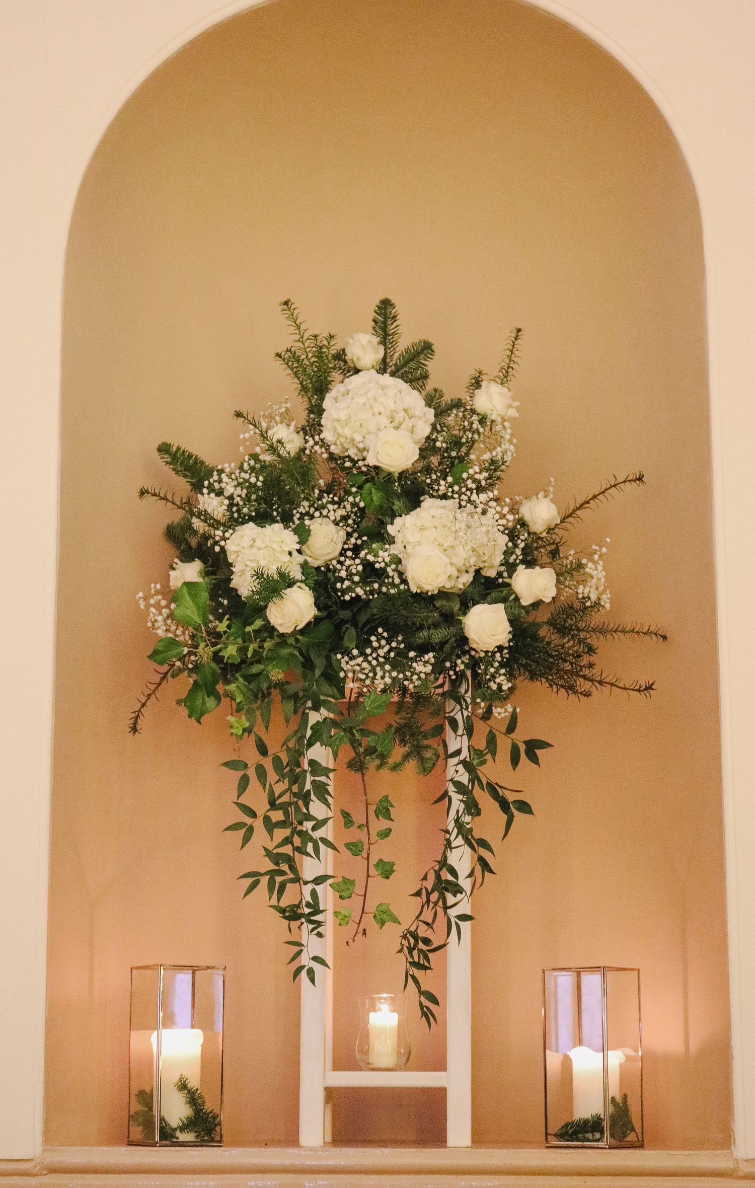 Elegant white floral arrangement with roses and baby's breath in an arched alcove flanked by glass-enclosed candles.