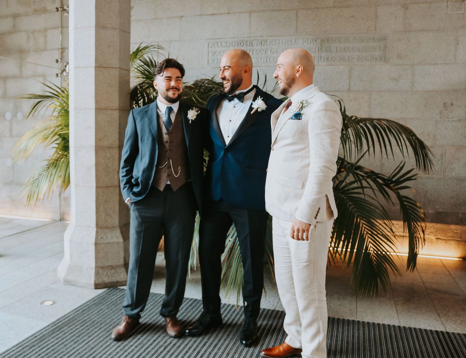 Three people in formal attire stand together at an elegant venue with palm plants in the background.