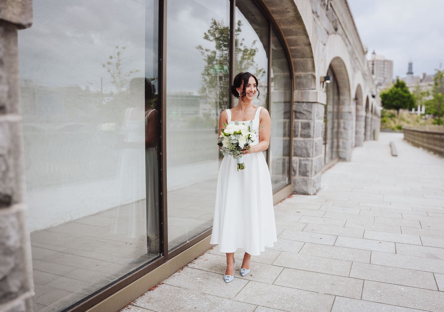 A person in a white midi dress holds a floral bouquet while standing by a stone archway near reflective windows.
