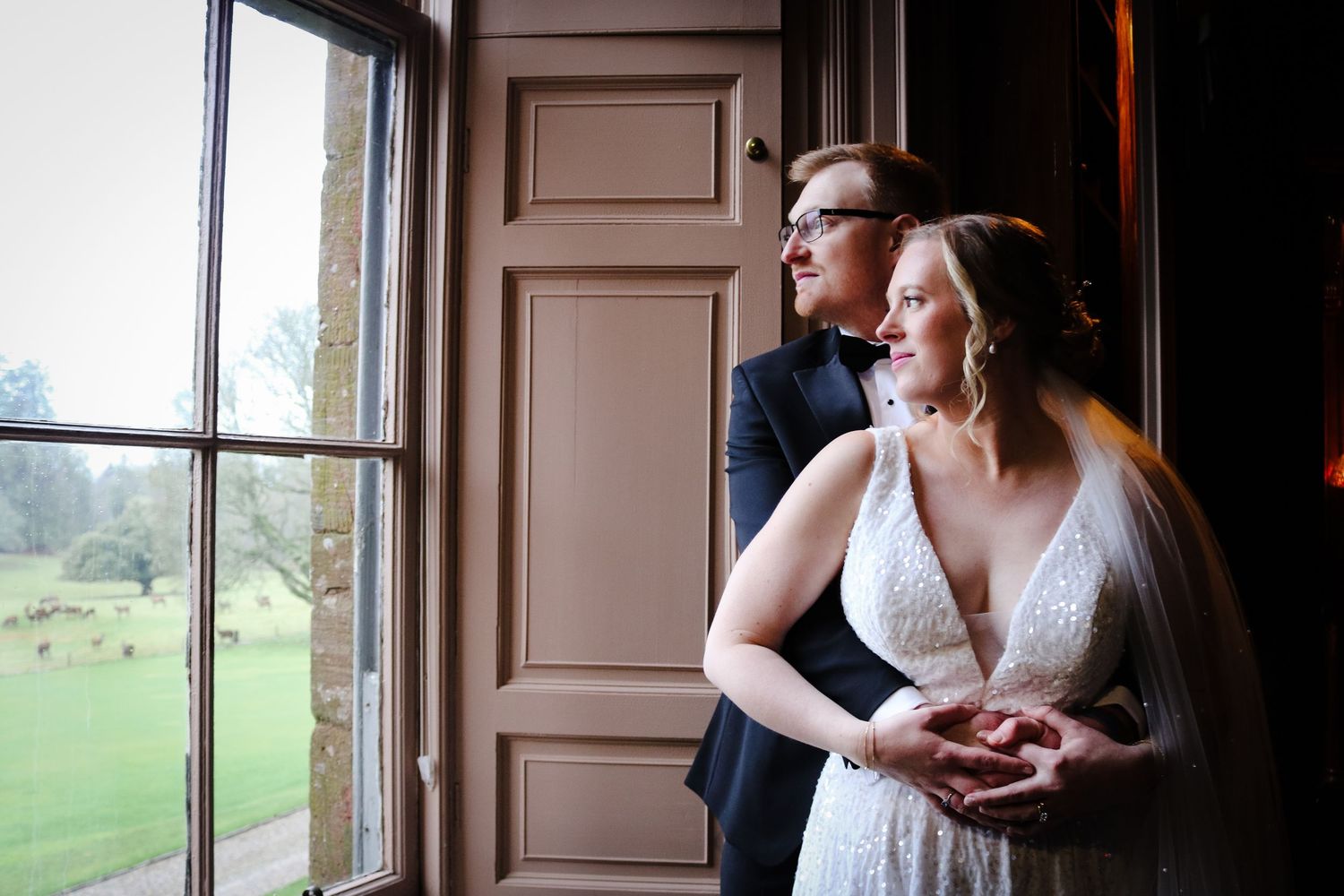 A couple in wedding attire gazes through a large window together in an elegant interior setting.