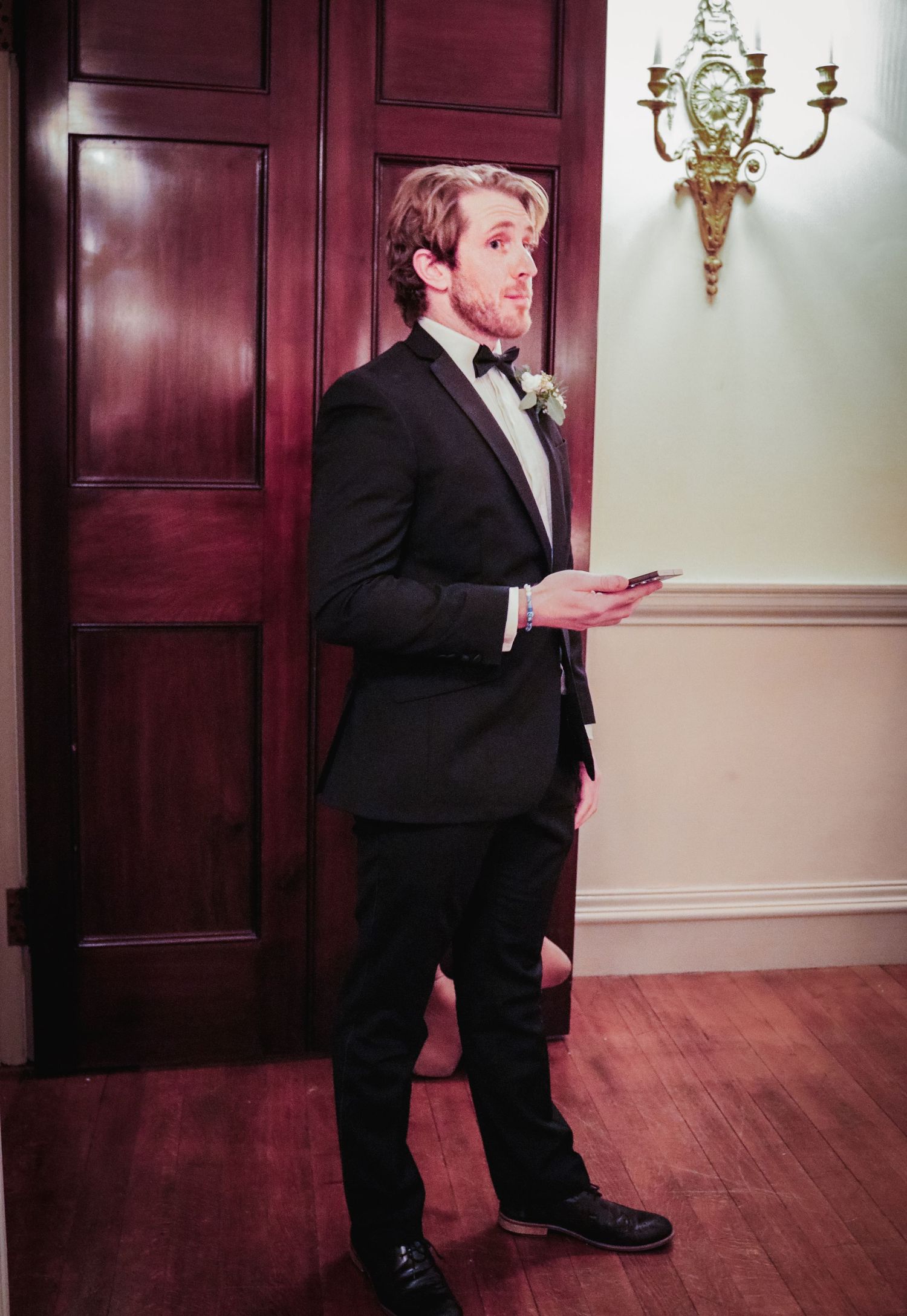 A person in a black tuxedo and bow tie stands in an elegant room with mahogany doors and a wall sconce.