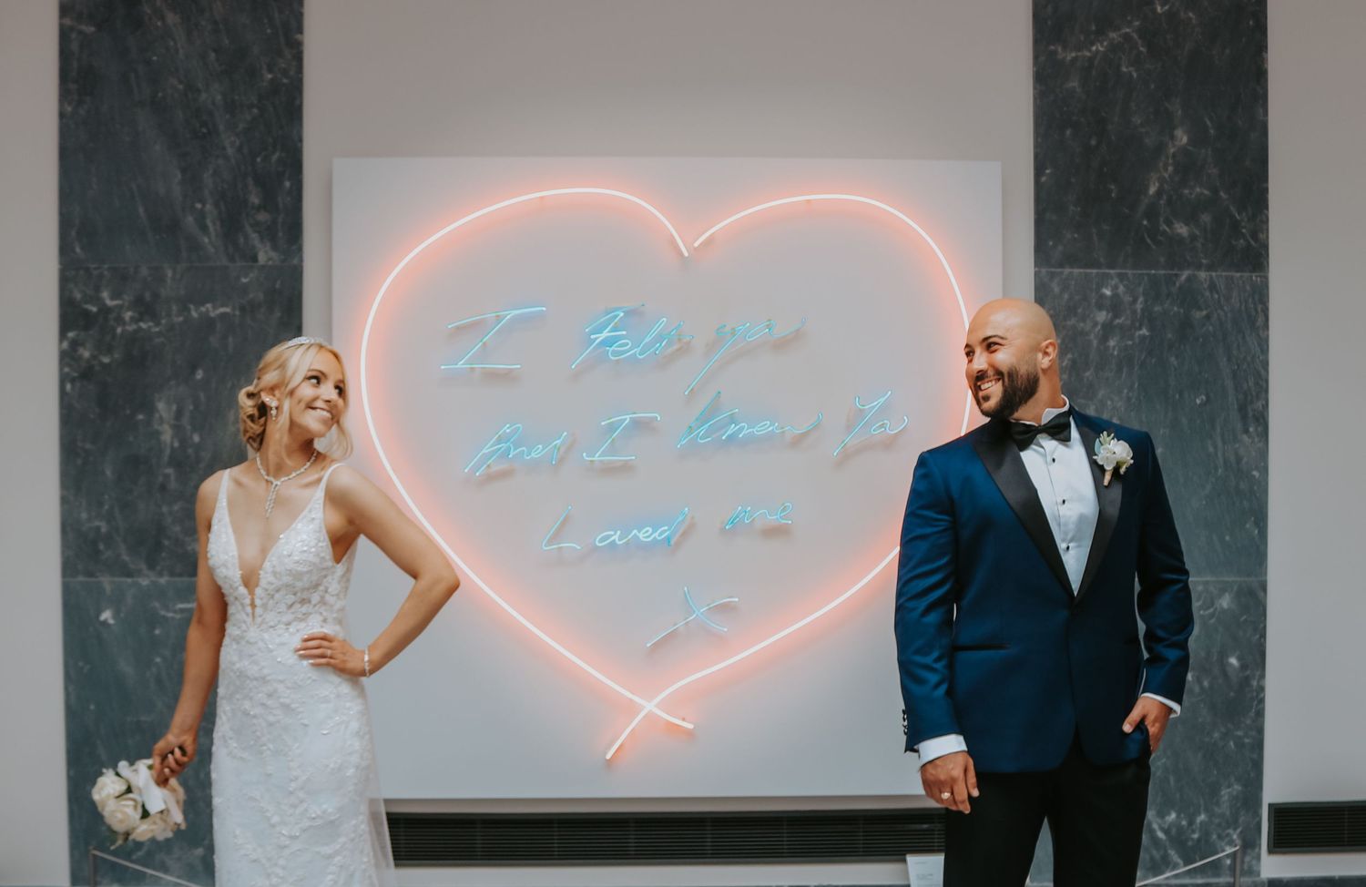 Wedding couple poses with pink neon heart sign on marble wall backdrop.