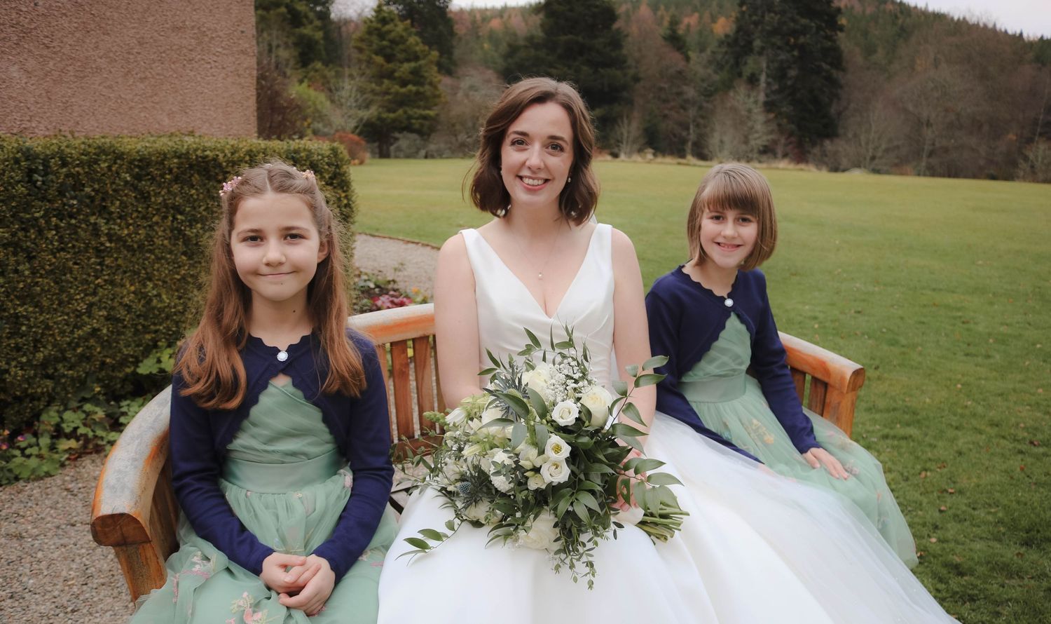 A bride in a white gown sits on a wooden bench with two flower girls wearing sage green and navy blue dresses outdoors.