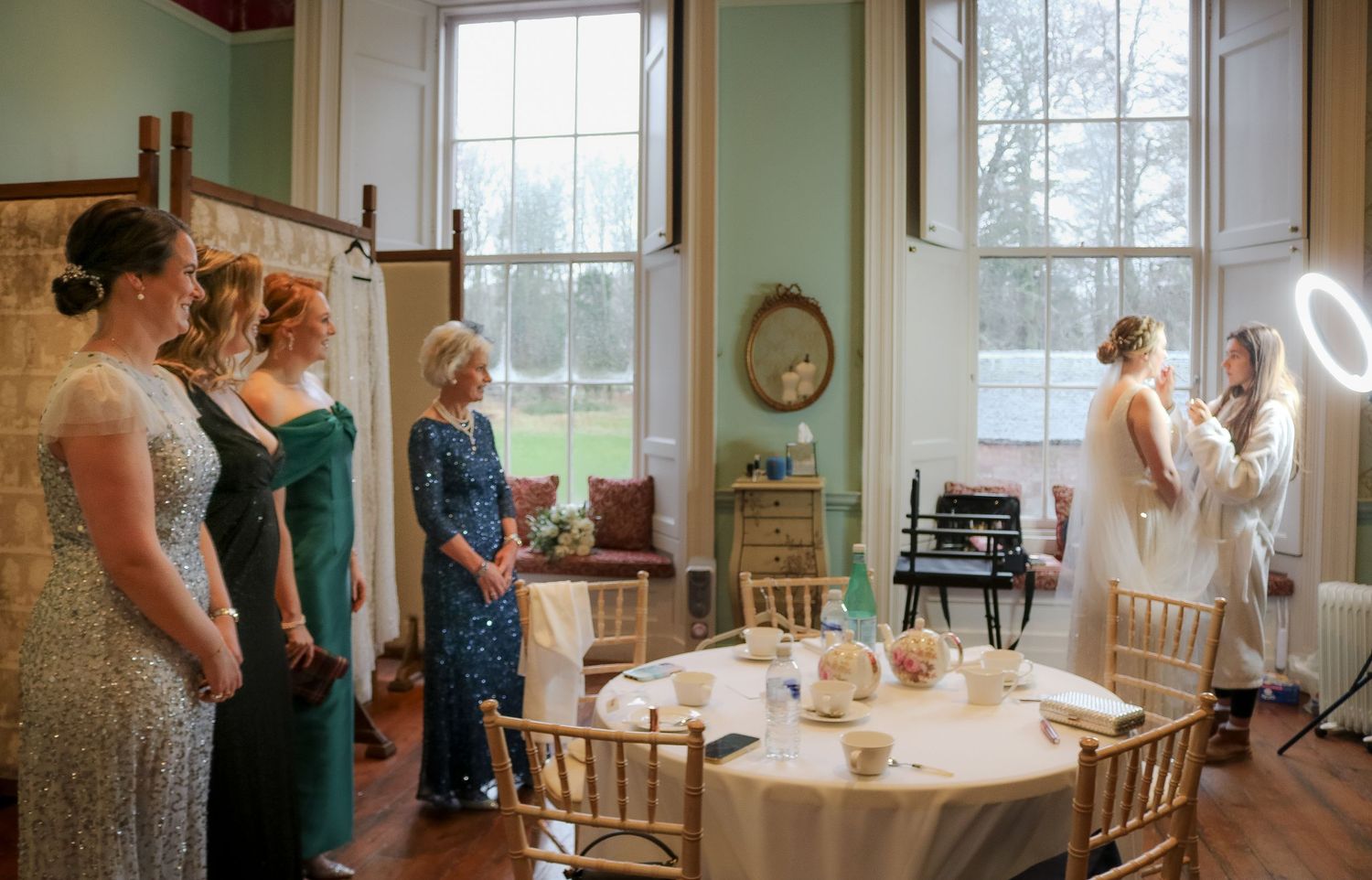 Bridesmaids gathered near elegantly set dining table in rustic interior with large windows and natural lighting.