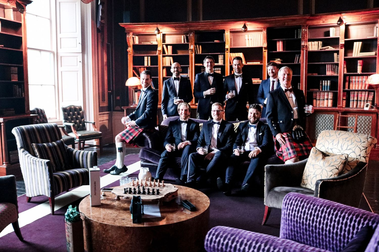 Elegant library room with plush purple furniture and chess set featuring groomsmen in dark suits gathered together.