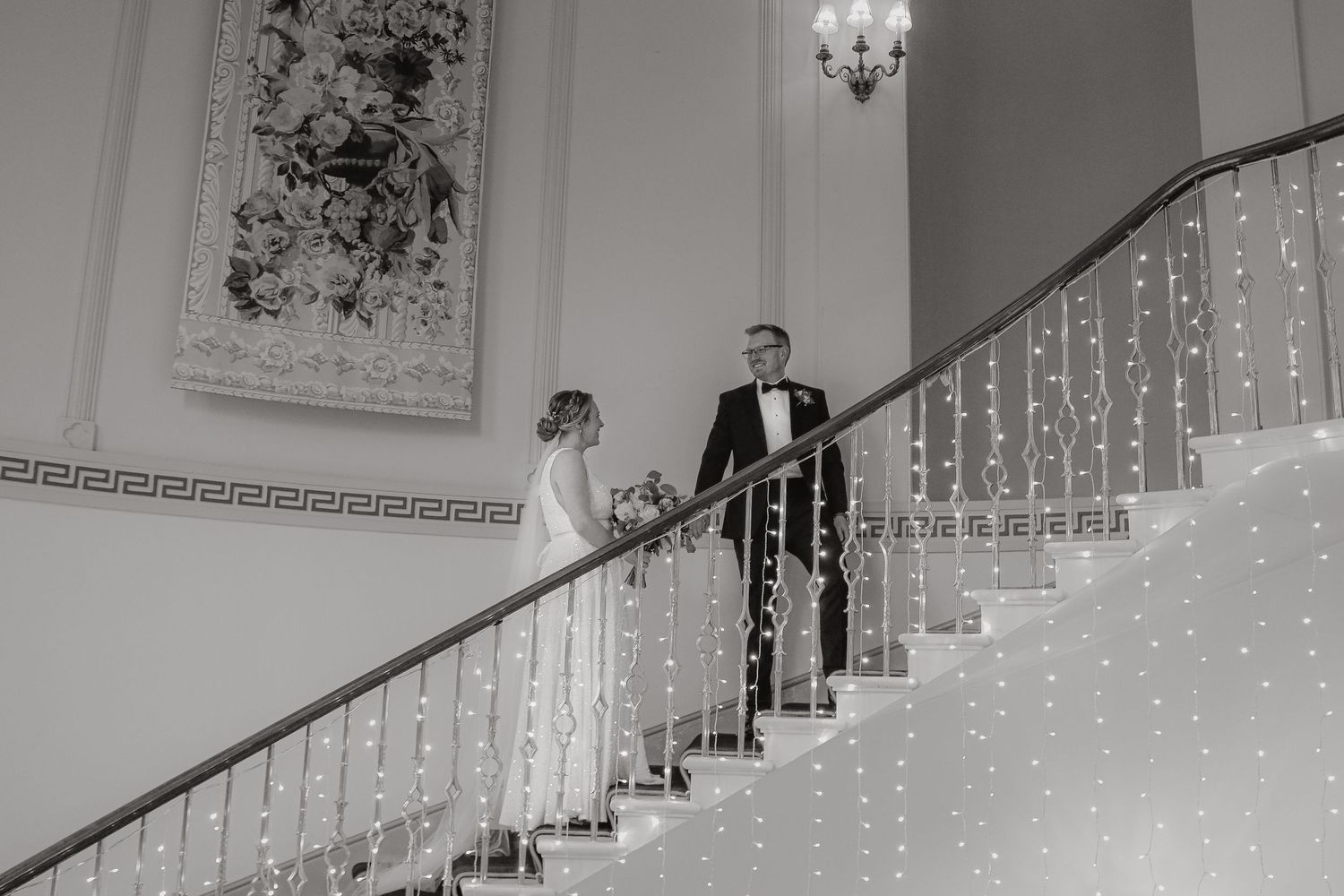 Two people in elegant formal wear stand on an ornate white staircase decorated with sparkling fairy lights.