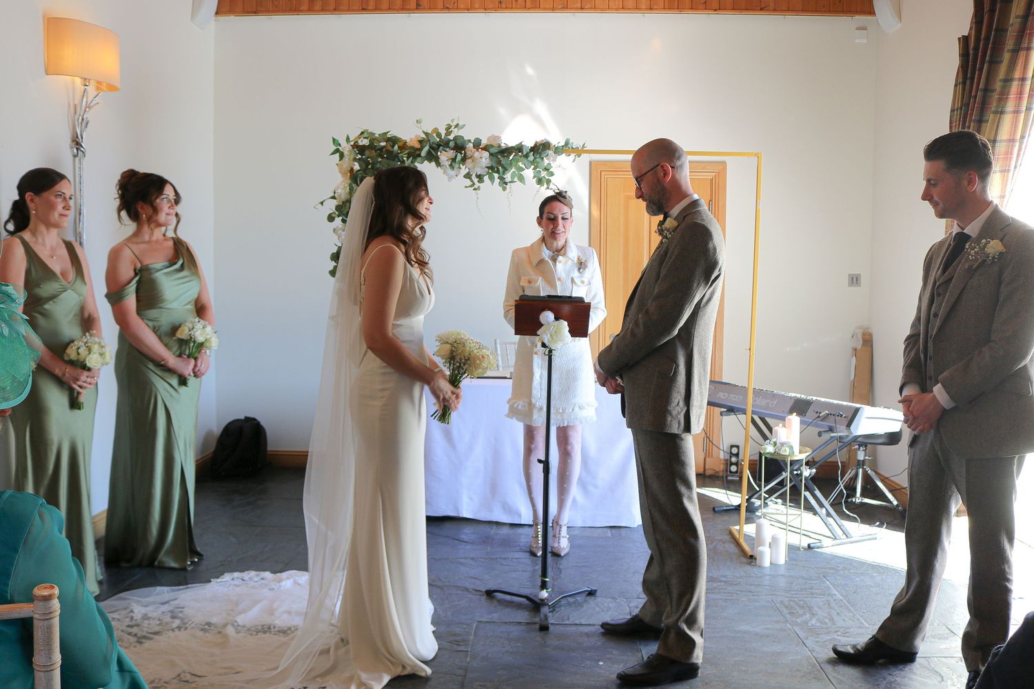 Wedding ceremony takes place in a bright indoor venue with mint green bridesmaid dresses and white floral decorations.