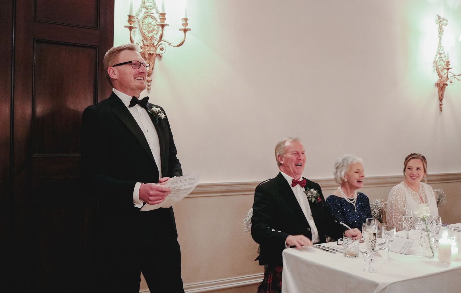 Someone delivers a speech at a formal reception while guests sit at a white-clothed table during what appears to be a celebration.