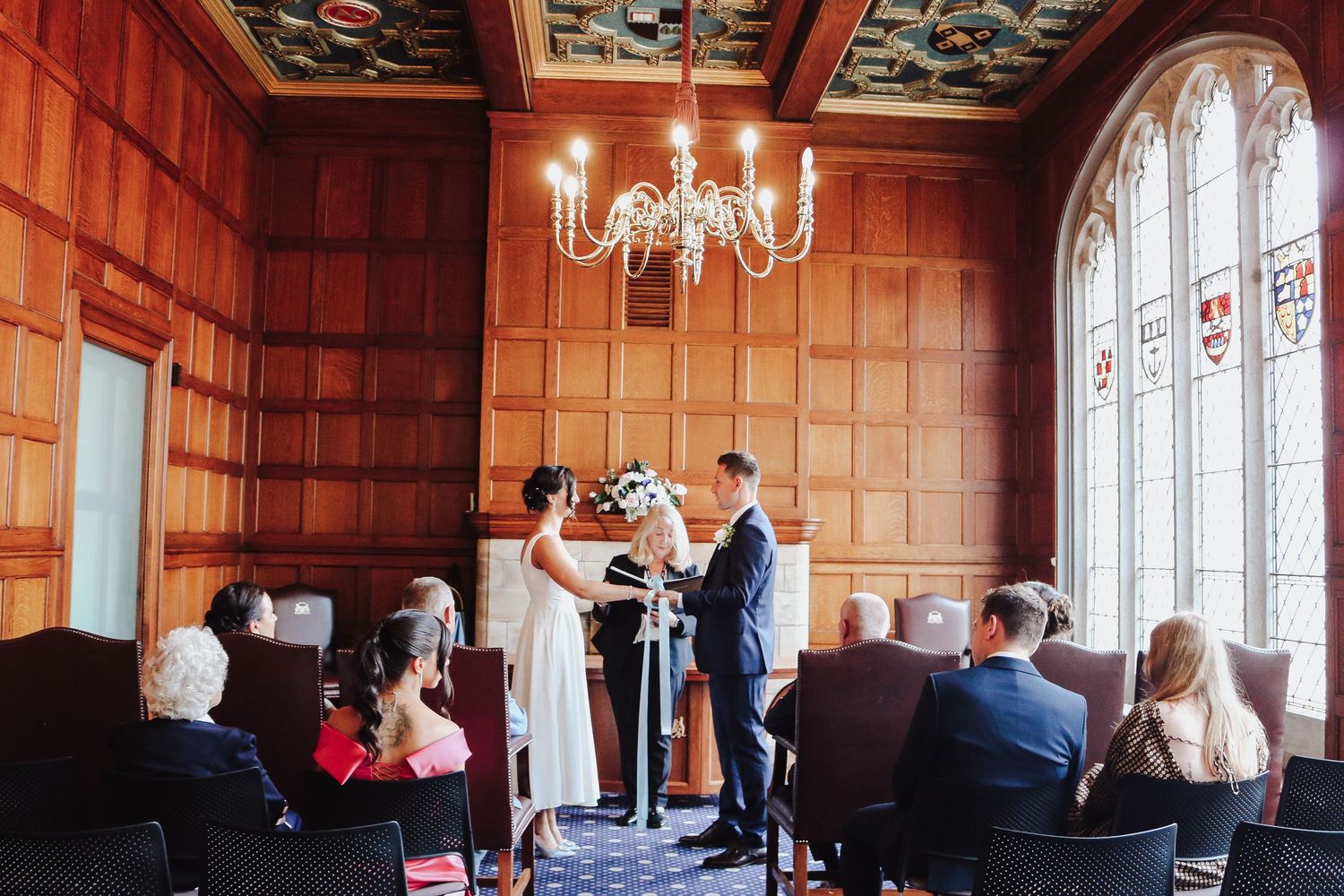 People gather in an elegant wood-paneled room with large windows and a chandelier for what appears to be a formal event.