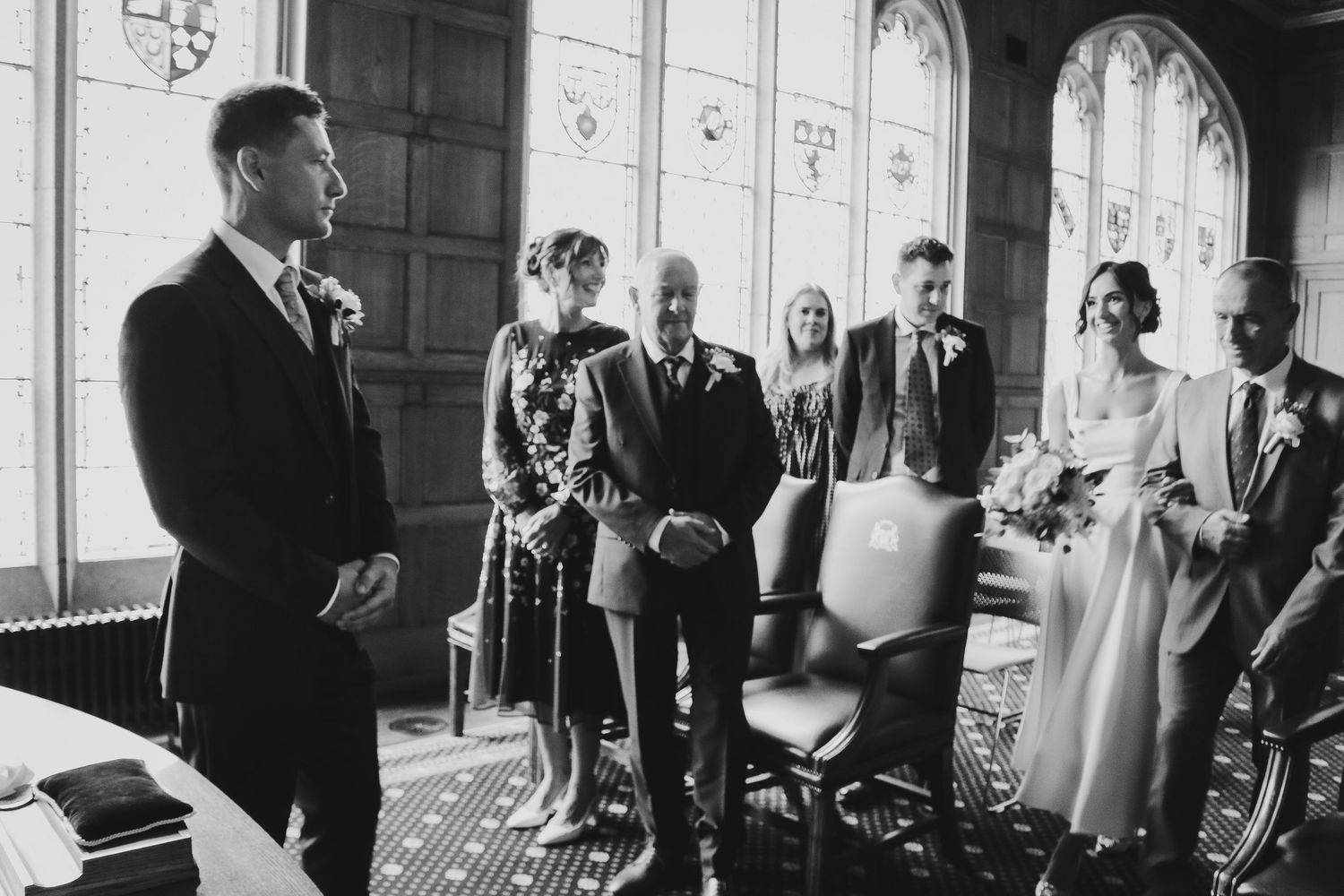 A black and white photograph showing a small wedding ceremony gathering in an elegant room with arched windows.